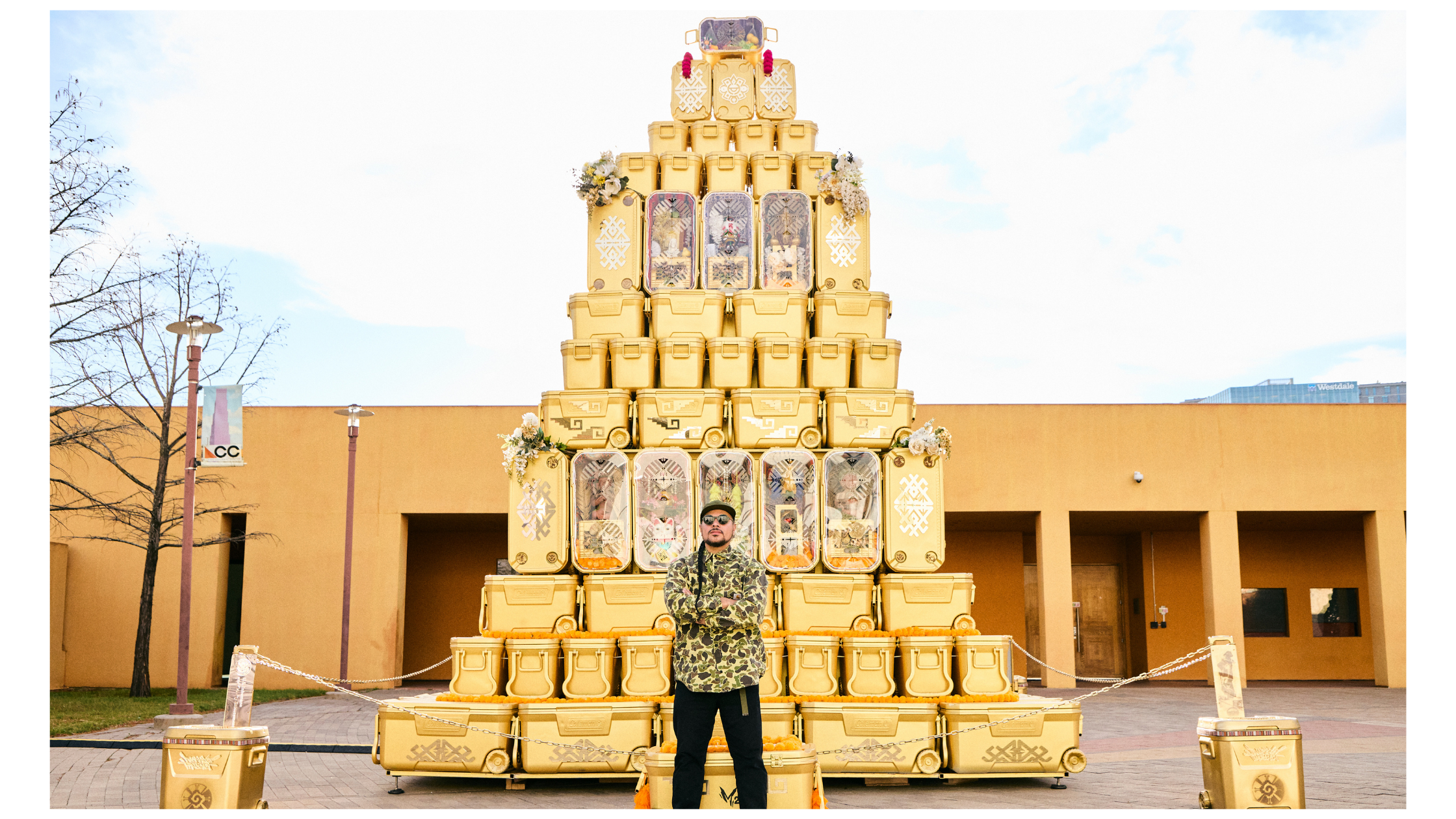 MARKA27 stands smiling in front of a large, ornate pyramid made of stacked golden ice coolers.