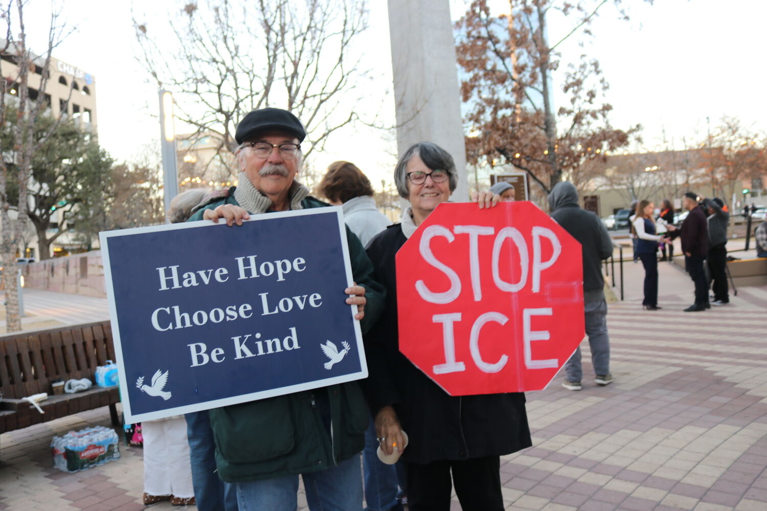 two people with signs. the person on the left holds a sign that says "have hope, choose love, be kind." the person on the right has a sign that says "stop ICE" in the shape of a stop sign.