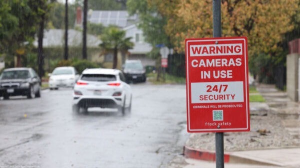 Rain falls on a residential street, where a red security sign warns "Cameras in Use 24/7 Security, Criminals Will Be Prosecuted, and flock safety." Cars drive down the wet road past houses, trees, sidewalks, and fences in the background.