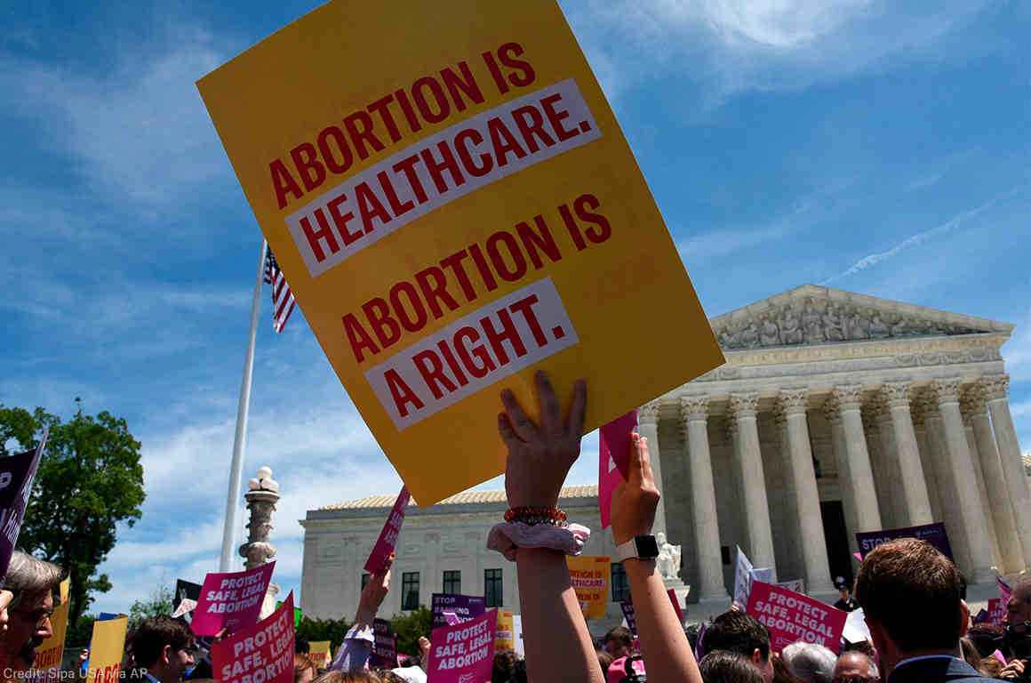 Image: A photo shows the US Supreme Courthouse in the background. Angled upwards, the camera captures the heads and arms of a large crowd of people. Directly in focus is an arm raising a sign that says "Abortion is healthcare. Abortion is a right."