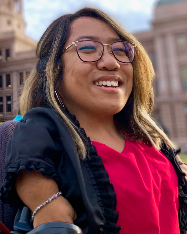 A disabled Mexican-American woman with medium-length hair. She is smiling to the camera, has glasses, and is wearing a black blazer with a pink blouse.