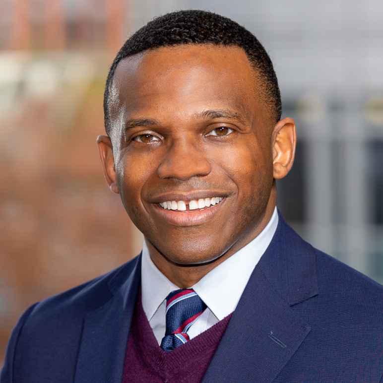 Photo of a man with short hair cut wearing a blue suit jacket, maroon vest, white shirt and tie.