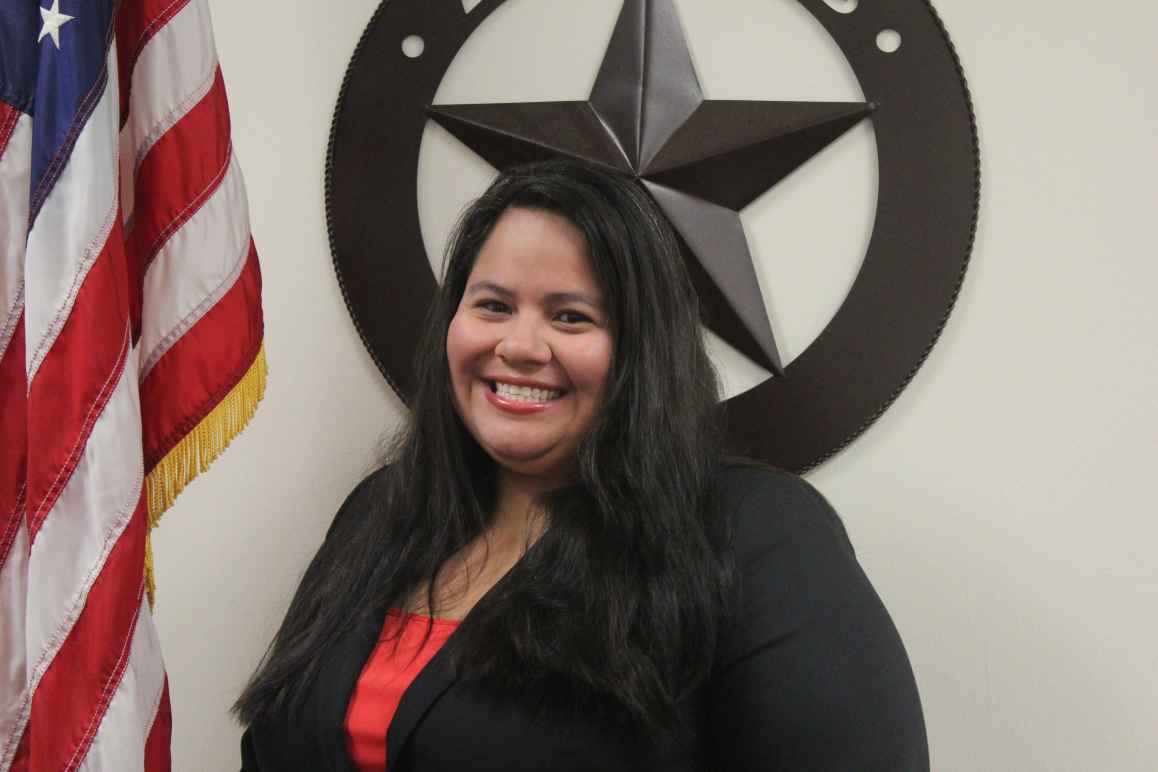 person with long dark hair, smiling, wearing a red blouse and a black jacket in from of a decorative lone star and American flag