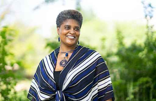 Photo of person with short dark hair, brown skin, smiling and wearing a white and black striped shawl in front of a green background