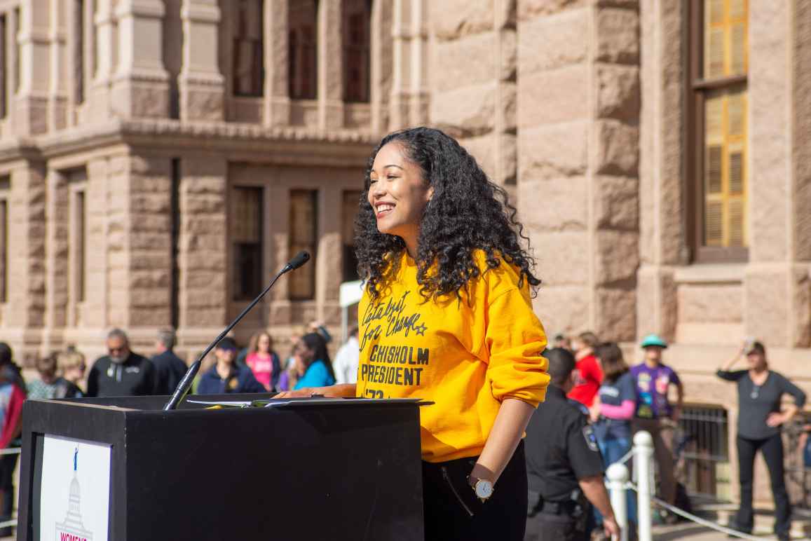 Photo of person standing at a podium smiling with curly dark hair, brown skin, wearing a yellow sweatshirt and black pants