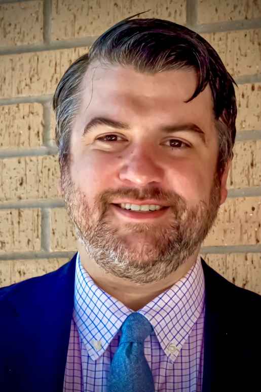 Person with short brown hair, plaid collared shirt, blue tie and dress coat