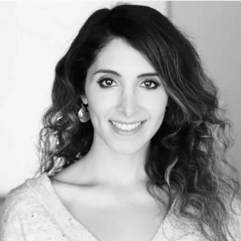 A black and white headshot image of a smiling woman with long, dark, wavy hair