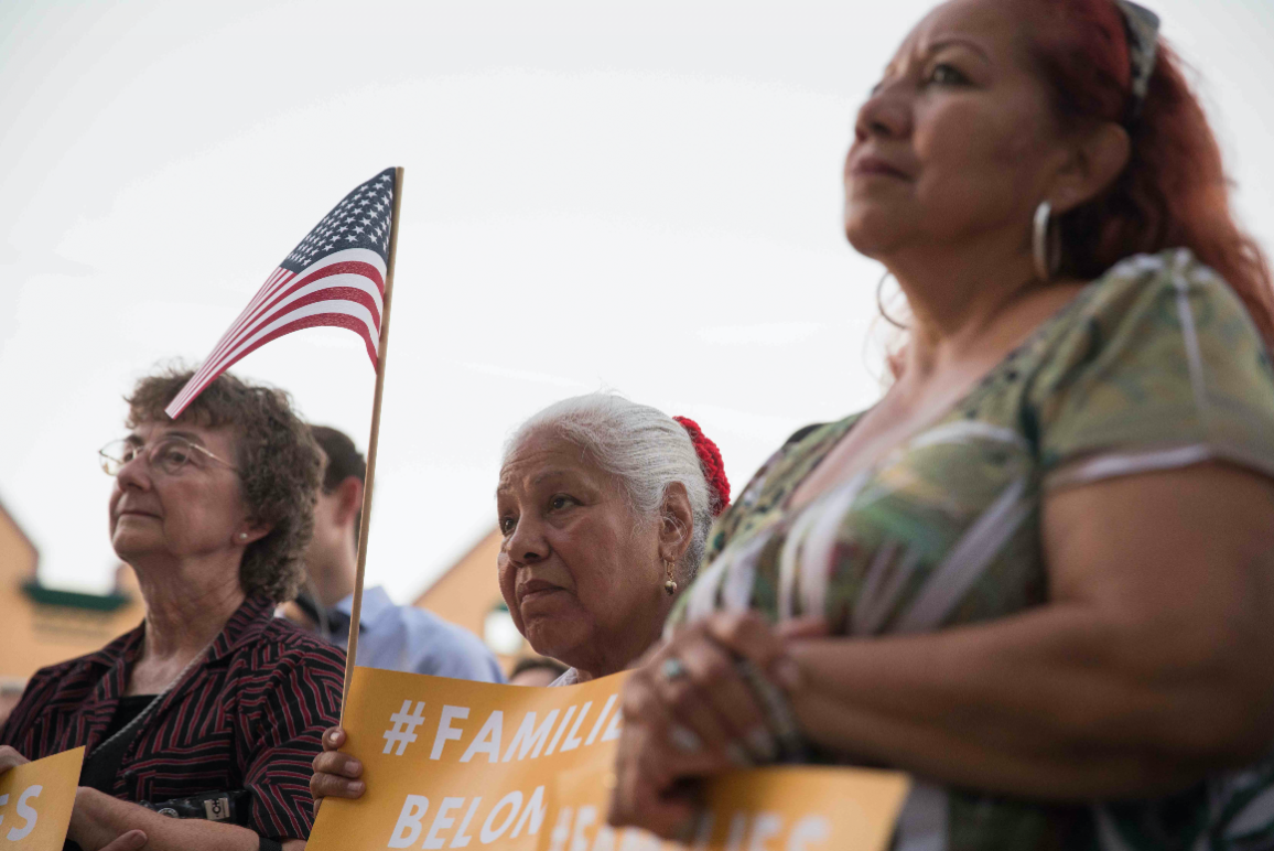 Image: Three women look on towards an unpictured speaker. They hold signs that say with the hashtag #FamiliesBelongTogether. One holds a U.S. flag up with her sign. 