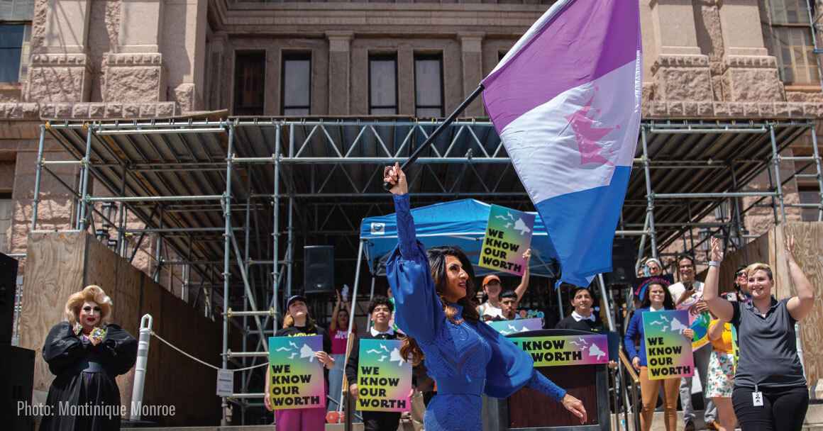 A drag queen raises a flag in front of supporters holding signs.