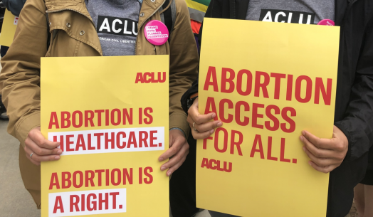 Two people are holding rally signs in support of reproductive freedom. On the left is someone holding a poster that reads "Abotion is healthcare. Aboriton is a right." On the right, a person holds a sign that reads "Abortino Access for All."