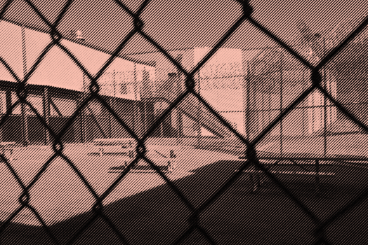 Image: A stylized photo shows an outdoor area of a detention center with some benches and tables. It is surrounded by barbed wire and concertina wire. The camera's view is partially obscured by the barbed wire fencing. 