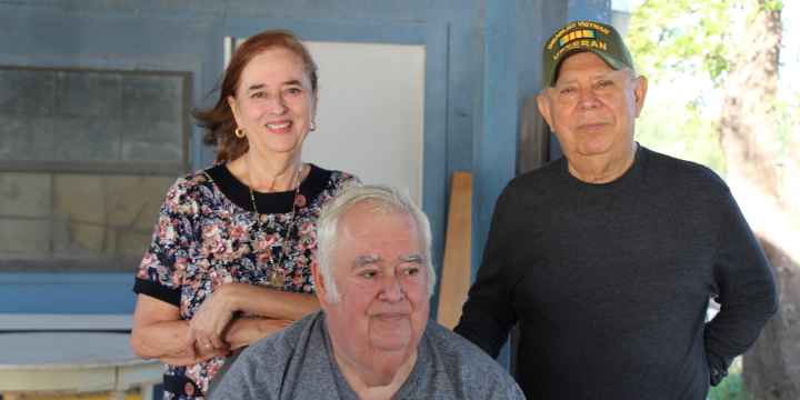 Cavozos family Image: A group of three pose for a photo outside of a wooden house, two standing and one sitting. The woman leans on the chair one man is sitting on. The man standing wears a baseball cap indicating he is a Vietnam War vet.