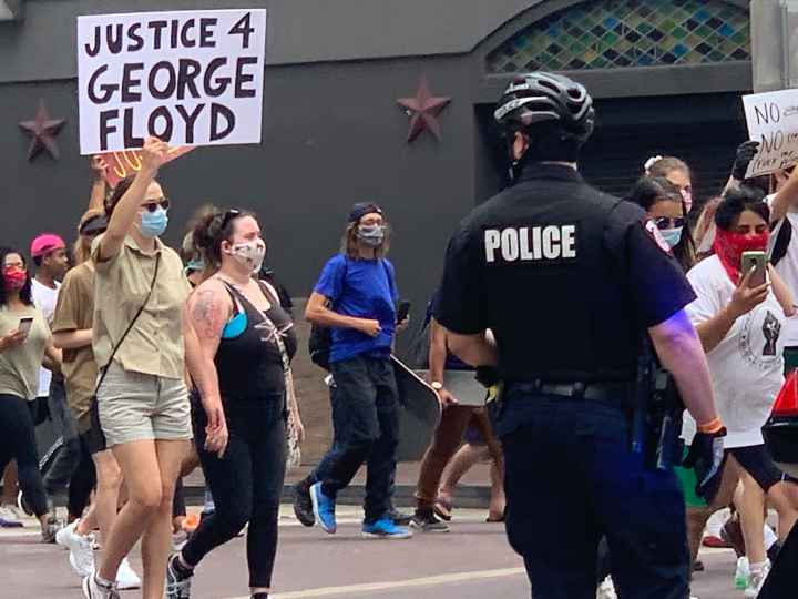 George Floyd protest Photo: A police officer looks on as a crowd of people march past at a downtown Houston intersection. In focus is a person holding up a sign that says 'Justice 4 George Floyd.'