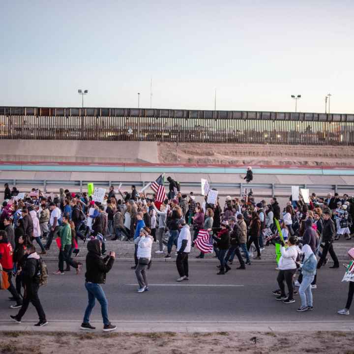 Photo: A large crowd streams by a a freeway and border fencing. They carry signs and american flags, walking in a general procession. 