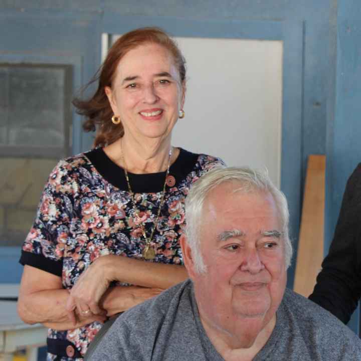 Cavozos family Image: A group of three pose for a photo outside of a wooden house, two standing and one sitting. The woman leans on the chair one man is sitting on. The man standing wears a baseball cap indicating he is a Vietnam War vet.