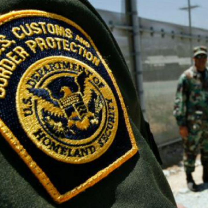 Photo: A Customs and Border Protection patch is worn on a green uniform. The CBP officer's arm takes us most of the photo, a fence and another CBP officer stand in the background.