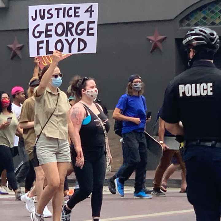 George Floyd protest Photo: A police officer looks on as a crowd of people march past at a downtown Houston intersection. In focus is a person holding up a sign that says 'Justice 4 George Floyd.'