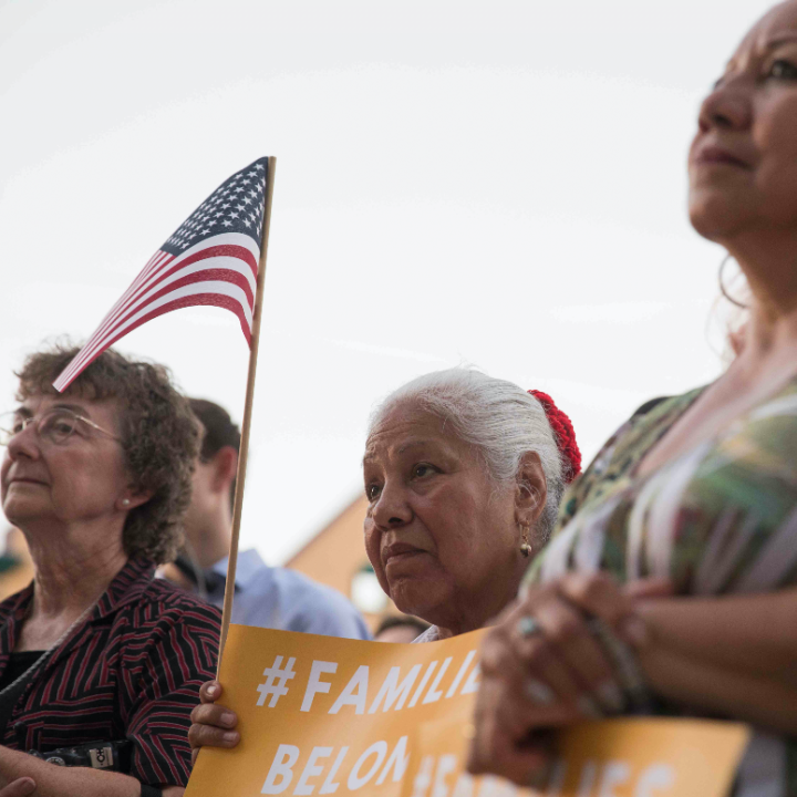 Image: Three women look on towards an unpictured speaker. They hold signs that say with the hashtag #FamiliesBelongTogether. One holds a U.S. flag up with her sign. 