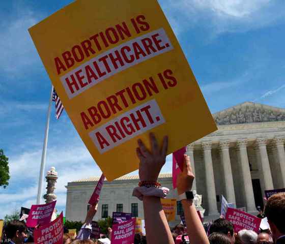 Image: A photo shows the US Supreme Courthouse in the background. Angled upwards, the camera captures the heads and arms of a large crowd of people. Directly in focus is an arm raising a sign that says "Abortion is healthcare. Abortion is a right."