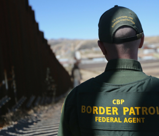 A photo shows a Border Patrol officer standing in the shadow of a border barrier. His back faces the camera and the barrier extends all the way to the horizon. 