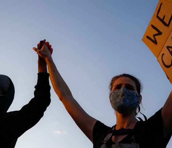 Photo: Two people wearing masks hold hands as they raise their arms. One hold a sign that says "We can't breathe"