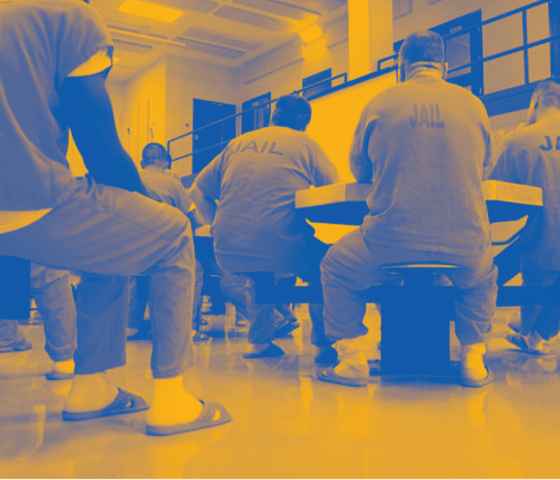 Image: A stylized photo shows men wearing uniforms that say "Jail" on their backs. They sit around tables in a carceral facility