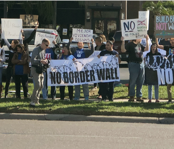 Photo: A crowd of people stand along a street in the Rio Grande Valley. Behind them is a restaurant and other businesses. They hold signs, one of which says "No Border Wall."