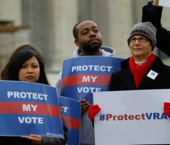 Photo: People gather, wearing cold-weather clothes, in front of the Supreme Court. They hold printed signs that say 'Protect My Vote" or "#ProtectVRA."